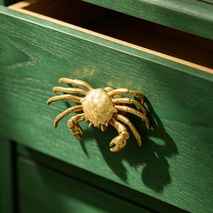 Puede incluir: Tirador de cajón dorado en forma de cangrejo montado en un cajón de madera verde. El cajón está parcialmente abierto, revelando el interior. El tirador de cangrejo tiene pinzas y patas detalladas, añadiendo un tema náutico al mueble.