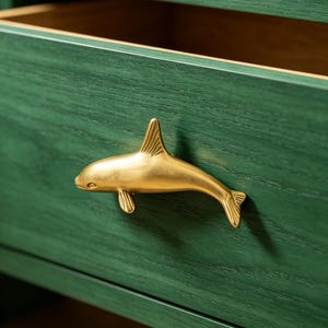 Könnte beinhalten: Ein goldfarbener Schubladengriff in Form eines Orca-Wals, montiert an einer dunkelgrünen Holzschubladenfront mit sichtbarer Holzmaserung. Die Schublade ist leicht geöffnet und gibt den Blick auf eine hellbraune Innenfläche im Schrank frei.