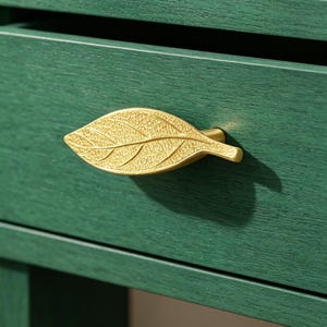 May include: Close-up of a green wooden drawer with a gold leaf-shaped handle. The handle is textured with detailed veining, adding a naturalistic touch to the furniture. The drawer is part of a larger piece of furniture.