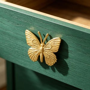 May include: A close-up of a green wooden drawer with a gold-colored butterfly-shaped handle. The butterfly handle is intricately detailed, and the drawer has a natural wood interior.