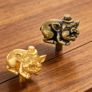 May include: Two metal cabinet knobs shaped like pigs wearing small top hats. One knob has a bright, polished gold finish, while the other features a darker, antique brass patina. Each pig is depicted in a reclining position with a smiling face.