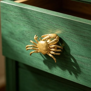 May include: A gold-toned metal cabinet knob shaped like a crab, featuring detailed legs and claws, mounted on a dark green wooden drawer front with a visible wood grain texture. The crab knob is centered on the drawer, casting a distinct shadow.