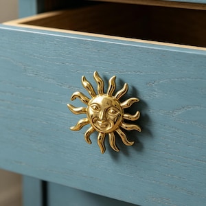 May include: A polished gold-toned metal drawer knob shaped like a sun with a human face, featuring radiating curved rays. The knob is mounted on a blue-painted wooden drawer front with a visible wood grain texture and an open top edge.