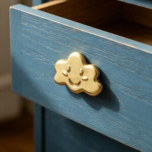 May include: A blue wooden drawer with a visible wood grain texture featuring a gold-toned metal cloud-shaped pull. The cloud pull has a stamped smiling face with eyes and a mouth. A second, smaller round gold knob is visible on the drawer below.