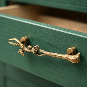 May include: A gold-colored drawer pull in the shape of a branch with a rose. The branch-shaped handle is mounted on a green wooden drawer. The drawer is open, revealing the interior.