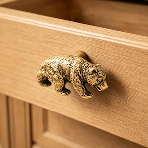 May include: A detailed, golden bear-shaped drawer pull mounted on a light-colored wooden drawer. The bear is sculpted with textured fur and stands out against the natural wood grain, adding a rustic touch to the furniture.