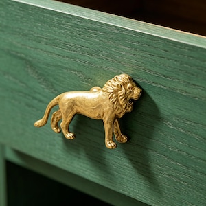 Puede incluir: Un tirador de cajón dorado en forma de león está montado en un cajón de madera verde. El león está de pie con la boca abierta. El cajón forma parte de un mueble más grande.