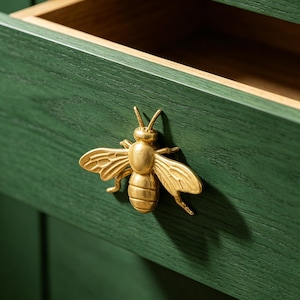 Puede incluir: Primer plano de un cajón de madera verde con un tirador dorado en forma de abeja. El cajón está abierto, revelando un interior marrón claro. El tirador de abeja añade un toque decorativo.