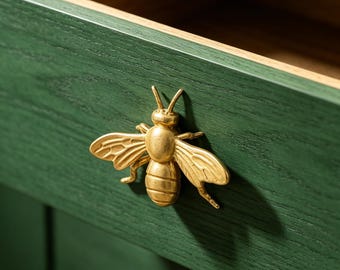 May include: Close-up of a green wooden drawer with a gold-colored bee-shaped handle. The drawer is open, revealing a light brown interior. The bee handle adds a decorative touch to the furniture.