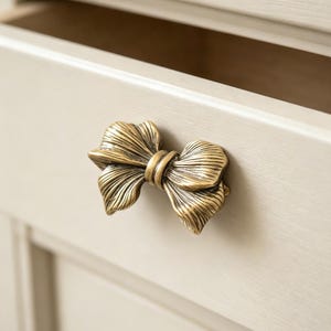 May include: Close-up of a gold-colored bow-shaped drawer pull. The drawer pull is attached to a light beige cabinet. The bow has detailed lines and a textured surface.