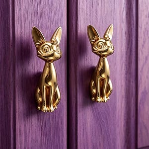 May include: Two polished gold-toned metal cabinet knobs shaped like sitting cats with pointed ears and detailed facial features. The cat-shaped hardware is mounted onto a purple wooden cabinet door with a visible vertical wood grain texture.