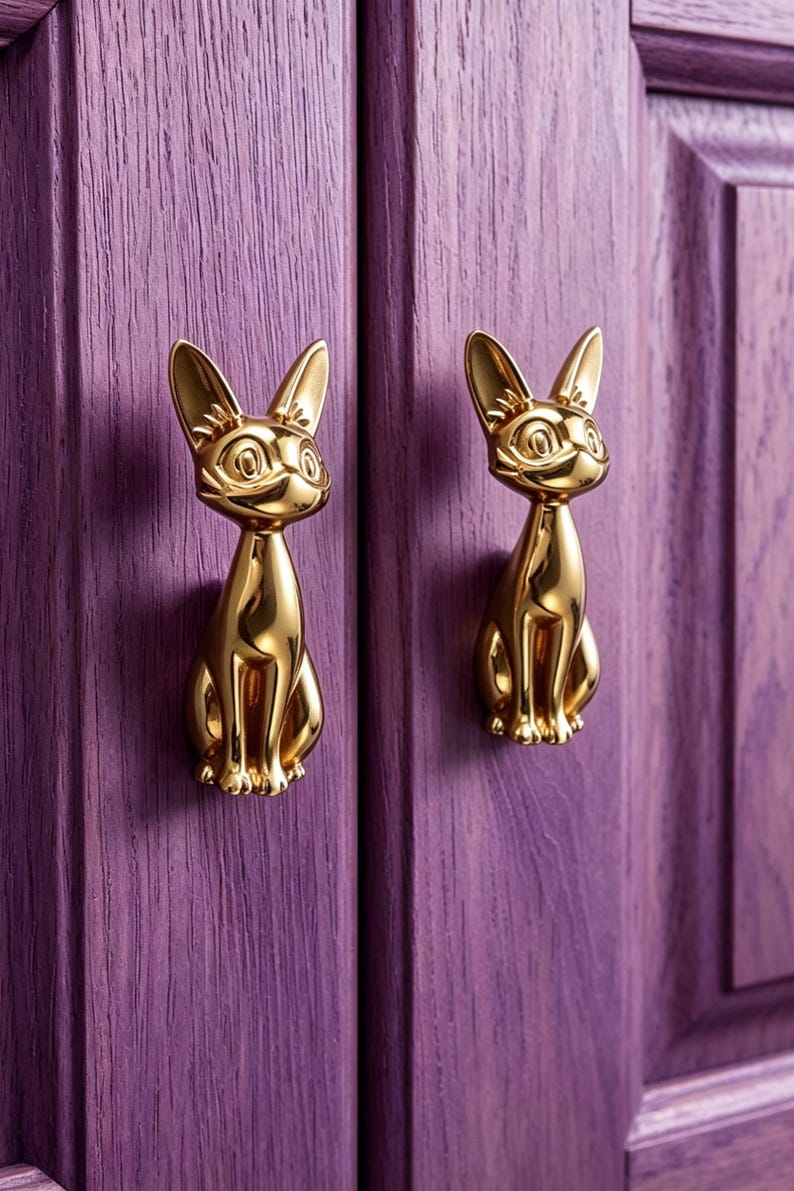 Op de afbeelding: Twee gepolijste goudkleurige metalen kastknoppen in de vorm van zittende katten met puntige oren en gedetailleerde gelaatstrekken. Het katvormige beslag is gemonteerd op een paarse houten kastdeur met een zichtbare verticale houtnerfstructuur.