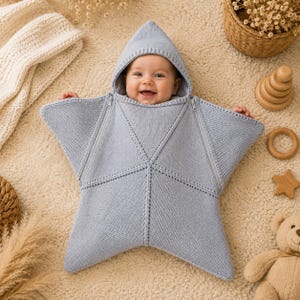 Può includere: Un sacco nanna per neonati a forma di stella lavorato a maglia in azzurro con cappuccio. Il sacco presenta un design con cucitura centrale e due cerniere diagonali che si estendono dal collo verso le punte esterne della stella.