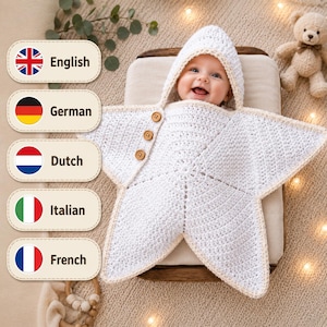 Könnte beinhalten: Eine weiße, gehäkelte Baby-Einschlagdecke in Form eines fünfzackigen Sterns mit Kapuze. Das Kleidungsstück ist mit drei kleinen braunen Holzknöpfen an einer Seite versehen und hat einen hellbeigen Rand an der Kapuze sowie an den Sternspitzen.