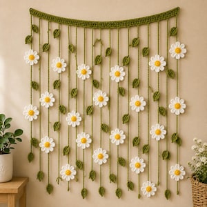 May include: A crochet wall hanging featuring a horizontal green band with multiple vertical green strings hanging down. Each string is decorated with small green crocheted leaves and white daisy flowers with yellow centers attached at various intervals.