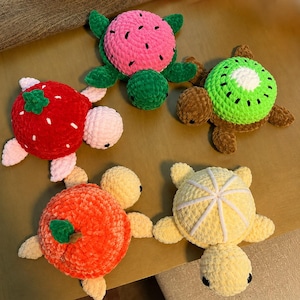 Puede incluir: Cinco tortugas de peluche hechas de hilo suave, cada una con un diseño de caparazón inspirado en frutas. La colección incluye una sandía rosa, un kiwi verde, una fresa roja, una naranja y un patrón de rodaja de cítrico amarillo pálido.