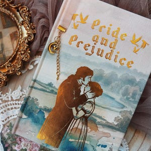 Op de afbeelding: Een hardcover boek getiteld Pride and Prejudice met een crèmekleurige stoffen kaft, voorzien van een gouden folie-illustratie van een omhelzend koppel, gouden tekst en vliegende vogels. Een gouden metalen kettingbladwijzer met maan- en hartbedel is bevestigd.
