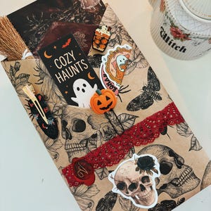 Op de afbeelding: Een rechthoekig pakket verpakt in beige papier met zwarte inktillustraties van schedels, motten en bloemen. Het bevat een rood kanten lint, een waszegel, een kleine bezem, een haarclip, een pompoenpaperclip en stickers van een geest en een schedel.