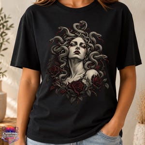 Peut inclure: Un t-shirt noir à manches courtes avec une impression graphique représentant une figure aux cheveux de serpents, rappelant Méduse, entourée de roses rouge foncé et de vignes épineuses. L'illustration utilise une palette de couleurs sourdes.