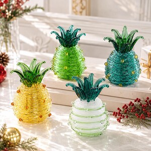May include: Four decorative pineapple-shaped glass ornaments featuring textured, coiled bodies and pointed green glass leaves. The set includes one yellow, one bright green, one teal, and one white pineapple, each accented with small glass beads.