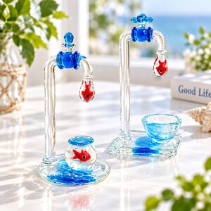 May include: Two clear glass sculptures featuring a blue faucet design. Each faucet has a red goldfish suspended in a glass drop. One sculpture includes a small round fishbowl with a red goldfish, while the other has a small blue glass bowl.
