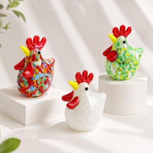 May include: Three glass chicken figurines featuring bright red combs, yellow beaks, and black eyes. One chicken has a multicolored speckled pattern, one is primarily green with yellow and white speckles, and one is solid white with a textured finish.