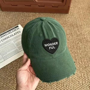 May include: A forest green baseball cap with a distressed brim and a black heart-shaped patch on the front. The patch features the white embroidered text WONDER FUL. The cap has a structured crown with ventilation eyelets and visible stitching.