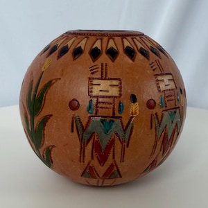 May include: A round, tan-colored gourd vessel featuring hand-carved and painted geometric figures. The surface displays stylized human-like forms in red, blue, and black, accented by green leaf motifs and a dark brown rim at the top opening.