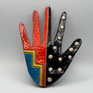 May include: A ceramic hand-shaped wall plaque featuring a split design. The left side is textured orange with a blue and yellow geometric pattern at the base. The right side is glossy black with raised white circular dots across the fingers and palm.
