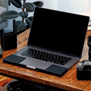 May include: A space gray laptop with a black screen and a black keyboard, featuring a black protective cover on the palm rest area. The laptop is positioned on a wooden surface next to a black camera and a black vertical stand.