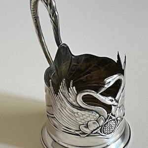 Op de afbeelding: Zilverkleurige metalen bekerhouder met een handvat. De bekerhouder heeft een sierlijk ontwerp van zwanen, bloemen en bladeren. Het handvat is gevormd als een gebogen steel met een getextureerde bovenkant. De bekerhouder is waarschijnlijk voor decoratieve doeleinden.