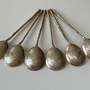 May include: Six silver spoons with ornate, engraved oval bowls and textured handles are arranged in a fan shape. The spoons have a diamond and floral pattern on the bowl.