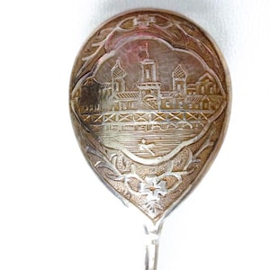 Könnte beinhalten: Ein silberfarbener Metalllöffel mit einer ovalen Laffe, die eine detaillierte Gravur eines Gebäudes mit Türmen und einer Brücke über Wasser zeigt. Der Griffbereich ist mit floralen Motiven und geschwungenen Mustern um das zentrale Bild verziert.
