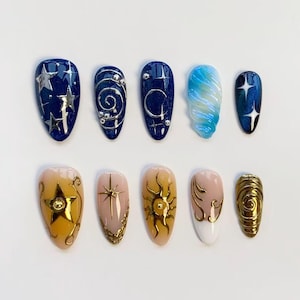 May include: A set of ten press-on nails featuring celestial designs. The top row includes dark blue glitter nails with silver stars, swirls, moons, and a blue marbled nail. The bottom row features tan and white nails with gold suns, stars, and swirls.