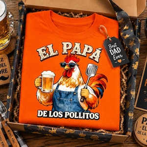 Può includere: Una t-shirt arancione con la grafica di un gallo che indossa occhiali da sole e un grembiule di jeans, mentre tiene in mano un boccale di birra e una spatola. Il testo recita EL PAPA DE LOS POLLITOS. La maglietta è in una scatola con un'etichetta BEST DAD EVER.