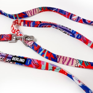Puede incluir: Una correa para mascota con un patrón abstracto multicolor en tonos rojo, azul, morado y blanco. La cinta de tela incluye una etiqueta rectangular negra con el texto blanco BERLINO y un mosquetón giratorio de metal plateado.