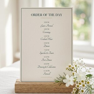 Peut inclure: Un panneau d'ordre du jour de mariage avec un fond crème et du texte noir. Le panneau est maintenu dans une base en bois et décoré de fleurs blanches. Le texte comprend le programme de la journée de mariage.