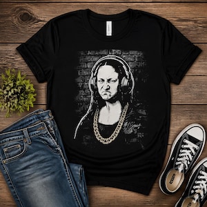 Könnte beinhalten: Ein schwarzes Kurzarm-T-Shirt mit einem Grafikdruck einer Person mit langem dunklem Haar, die große Over-Ear-Kopfhörer und eine dicke goldfarbene Kette trägt. Die Figur hat einen strengen Gesichtsausdruck und ist vor einer Ziegelwand abgebildet.