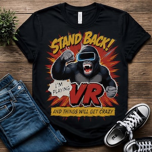 May include: A black t-shirt featuring a graphic of a gorilla wearing a virtual reality headset. The text reads STAND BACK! I'M PLAYING VR AND THINGS WILL GET CRAZY. The design includes a red and yellow explosion background behind the gorilla.