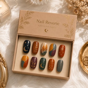 May include: A set of ten press-on nails in a tan cardboard box labelled Nail Reverie Press-On Nails. The nails feature various designs including gold moons, stars, autumn trees, and botanical patterns in shades of navy, orange, green, and purple.