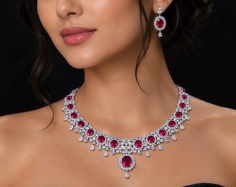 Puede incluir: Un conjunto de collar y pendientes a juego en tono plateado con piedras ovaladas de color rojo vibrante rodeadas de pequeños cristales blancos transparentes. El collar tiene un colgante central de piedra roja ovalada con varias piedras rojas más pequeñas a lo largo de la cadena.
