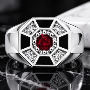 Puede incluir: Un anillo de tono plateado con una piedra roja central de corte redondo rodeada por pequeñas esferas plateadas. La cara del anillo tiene un diseño geométrico octogonal con cuatro piedras claras y cuatro secciones triangulares de esmalte negro.