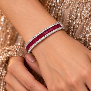May include: A silver-toned hinged bangle bracelet featuring a central row of rectangular, deep red baguette-cut stones flanked by two parallel rows of small, round, clear, brilliant-cut stones set in a metallic frame.