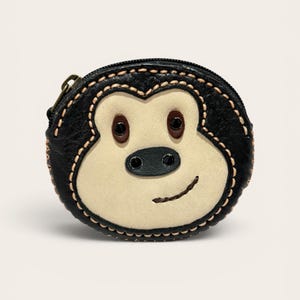 May include: A round coin purse shaped like a monkey face, crafted from black and cream-colored leather. The design features a black zipper closure, dark brown eyes, a black nose, and a stitched mouth, with visible tan stitching along the edges.