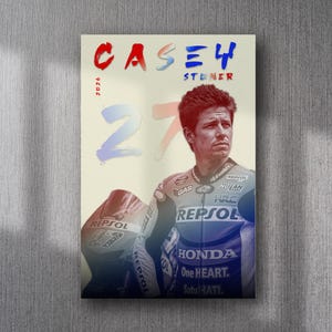 Può includere: Poster con un ritratto di Casey Stoner in tenuta da corsa, con la scritta "CASEY 4 STONER 27" in rosso e blu. Il poster include i loghi Honda e Repsol e un casco da corsa.