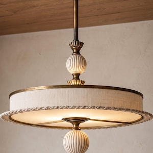 May include: A ceiling-mounted pendant light fixture featuring a circular drum shade in off-white fabric with a braided rope trim. The central stem and decorative accents are finished in antique brass, including two fluted, spherical ceramic finials.
