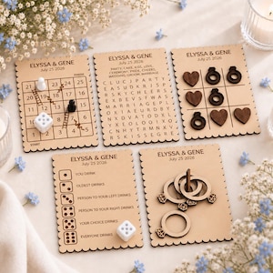 May include: Five rectangular wooden game boards with scalloped edges, personalized with 'ELYSSA & GENE July 25 2026'. Boards feature a numbered ladder game, a word search, a tic-tac-toe grid with heart and ring tokens, a dice drinking game, and a ring toss.