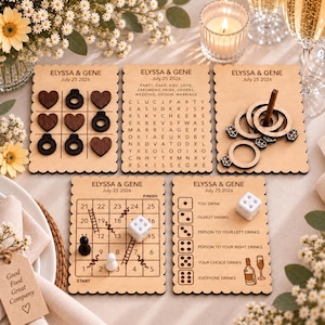 May include: Five rectangular wooden game boards with scalloped edges, each engraved with ELYSSA & GENE July 25 2026. Includes a tic-tac-toe board with dark heart and ring pieces, a word search, a ring toss, a numbered board game, and a dice drinking game.