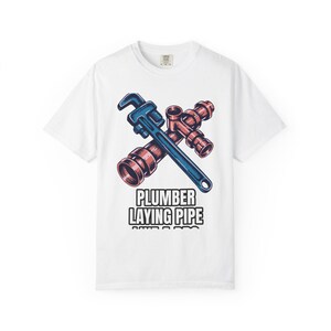 Puede incluir: Camiseta blanca con un diseño de una llave inglesa azul y tuberías de cobre. El texto "PLUMBER LAYING PIPE" está impreso debajo. La camiseta es de cuello redondo.