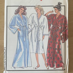 Op de afbeelding: Een vintage naaipatroonpakket met illustraties van drie verschillende badjassen. Het pakket is gelabeld met "Style", maat "XL" en nummer "1427". De badjassen zijn blauw, wit en rood met bloemmotieven.